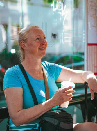 Photo for Young beautiful caucasian blonde woman with a braided braid sits on a bus stop bench outdoors, holding a suitcase handle with one hand and a paper glass of water and a straw with a joyfully happy smile on her face, waiting for an arriving tram, close - Royalty Free Image