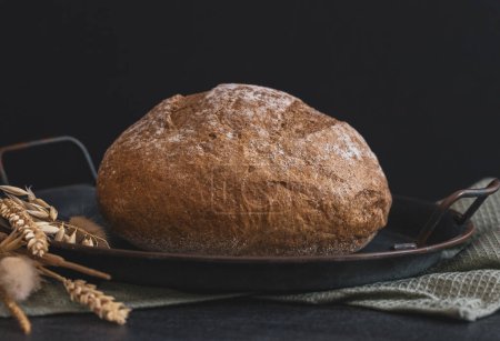 Foto de One round rye bread in a vintage metal plate with a kitchen napkin and ears lie on a black stone background, close-up side view.Bread baking concept. - Imagen libre de derechos