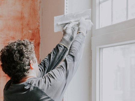 Téléchargez les photos : A caucasian young guy a plasterer in a gray T-shirt with sleeves and curly brown hair levels fresh putty with a spatula on a window opening, side view close-up. The concept of repairing apartments and - en image libre de droit