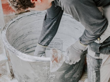 Téléchargez les photos : Caucasian young guy-plasterer in a gray T-shirt with long sleeves and curly brown hair, standing in an inclination, is picking up fresh putty from a large black basin on the floor, close-up side view - en image libre de droit
