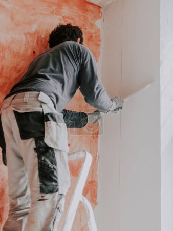 Téléchargez les photos : A caucasian young guy, a plasterer in a gray T-shirt with sleeves, work pants and curly brown hair, smoothes fresh putty with a large spatula on the upper wall near the ceiling, pulling down and - en image libre de droit