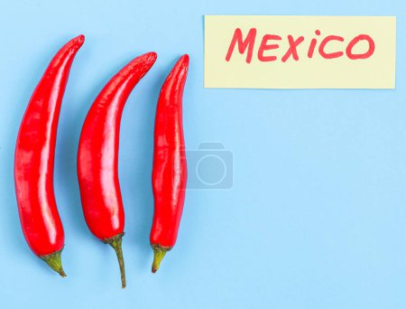 Foto de Tres piezas de chiles rojos picantes con una nota mexicana amarilla sobre un fondo azul claro con espacio para copiar, plano de primer plano. Concepto de Cinco de Mayo, minimalismo, espacios en blanco. - Imagen libre de derechos
