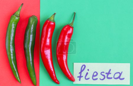 Foto de Four hot green and red chili peppers with the inscription on paper: fiesta and copy-space lie on a red-green background, flat close-up. Cinco de Mayo concept, minimalism, blanks. - Imagen libre de derechos