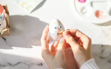 Photo for Hands of caucasian teenage girl in white turtleneck holding easter bunny egg and pasting sticker bow while sitting at marble table with palette and stickers, flat lay closeup. The concept of crafts, needlework, at home, diy,children art,artisanal,eas - Royalty Free Image