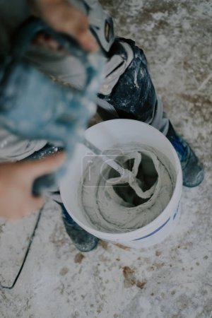 Photo for Young caucasian man unrecognizable whisking white wet putty with a construction mixer in a white small bucket, closeup top view with selective focus. The concept of home renovation, construction work. - Royalty Free Image