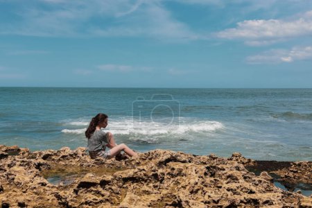 Téléchargez les photos : Une adolescente brune caucasienne en short et chemisier est assise sur un rivage rocheux, regardant de près la mer du Nord à Etretat, en France. Concept des personnes et de la nature. - en image libre de droit