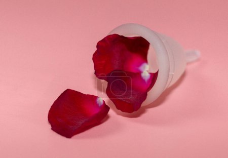 Foto de Una copa menstrual de silicona con pétalos de rosa roja saliendo de ella se encuentra sobre un fondo rosa, vista lateral de primer plano. Concepto de salud femenina. - Imagen libre de derechos
