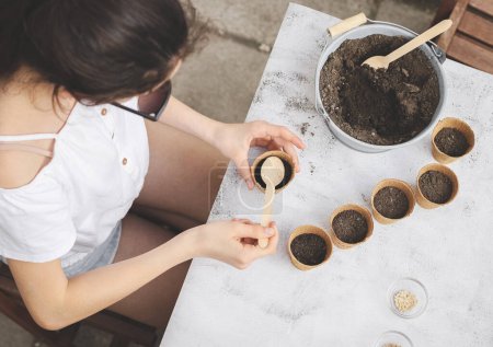 Photo for Caucasian teenage girl sits at a table on the street in the backyard of the house, holding a glass in cardboard and pouring soil into it with a wooden spoon on a light cement background, flat lay close-up. Concept gardening, sowing seeds, seedlings, - Royalty Free Image
