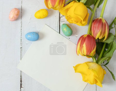 Foto de Rosas amarillas, tulipanes ardientes, huevos de pascua de mármol y un sobre blanco vacío yacen sobre una mesa de madera blanca con espacio para copiar, plano ponen de cerca. - Imagen libre de derechos