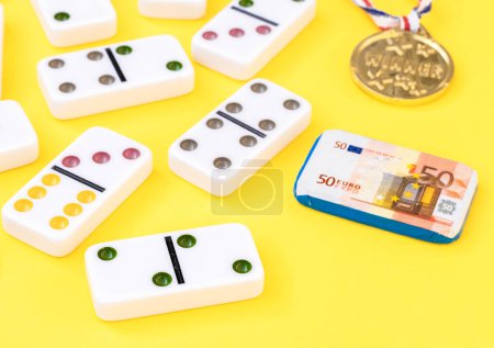 Téléchargez les photos : White dominoes,winner's medal and chocolate euros on yellow background,close-up side view with depth of field.Board game concept. - en image libre de droit
