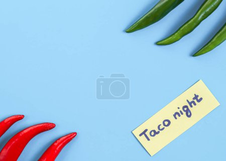 Foto de Los chiles verdes y rojos picantes se encuentran en los lados con una nota nocturna de taco amarillo sobre un fondo azul claro con espacio para copiar, de primer plano. Concepto de Cinco de Mayo, minimalismo, espacios en blanco. - Imagen libre de derechos