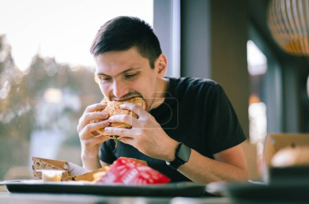 Foto de Retrato de un joven guapo morena caucásica con una camiseta negra se sienta en una mesa cerca de la ventana en un restaurante y come apetitosamente una hamburguesa, mordiéndola, vista lateral de cerca. - Imagen libre de derechos