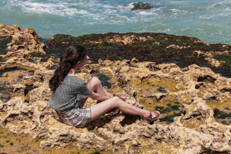 Téléchargez les photos : Une adolescente brune caucasienne en short et chemisier est assise sur un rivage rocheux, regardant de près la mer du Nord à Etretat, en France. Concept des personnes et de la nature. - en image libre de droit