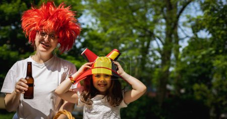 Téléchargez les photos : Portrait de belle et heureuse maman caucasienne en perruque rouge et petite fille portant un chapeau drapeau belge célébrant le jour du belgique au pique-nique dans le parc de la ville le jour ensoleillé d'été, vue latérale rapprochée - en image libre de droit