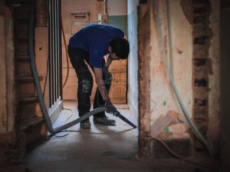 Caucasian young professional janitor in uniform vacuums debris with a construction vacuum cleaner in the aisle near the landing in an old and dirty house preparing it for repairs standing in a dark