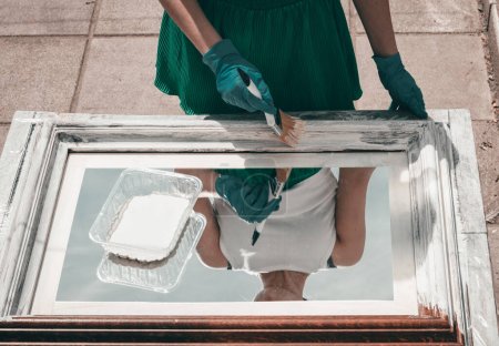 Photo for One young Caucasian unrecognizable girl in green shorts and gloves paints a mirror frame with white paint with a wide brush, reflecting in it while standing in the backyard of the house on a summer day, close-up top view. - Royalty Free Image
