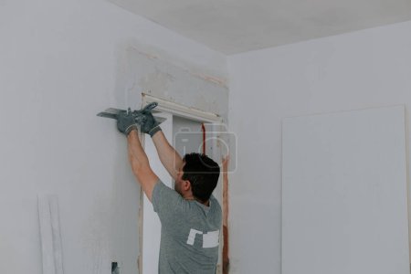A young Caucasian recognizable brunette in a gray uniform and gloves covers the construction grid with white putty with a spatula, standing half-turned on a stepladder in the doorway, close-up view from below with selective focus. The concept of home
