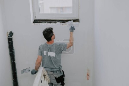 Photo for A handsome young Caucasian man in a gray uniform and gloves, standing from his back, plasters a window opening on the landing of the second floor with a small spatula, close-up side view. Construction concept. - Royalty Free Image