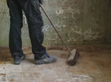 Photo for One young unrecognizable man in a uniform sweeps garbage with a broom from a dirty tiled floor in an old house preparing for renovation, side view close-up. - Royalty Free Image