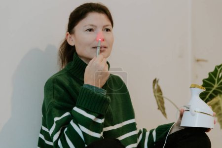 Photo for One beautiful Caucasian brunette girl with collected hair in a green sweater treats the right nasal passage with an apparatus with infrared light, sitting on a bed against a white wall, close-up view - Royalty Free Image