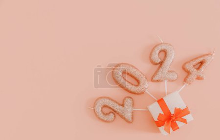Photo for Shiny candles number 2024 with one small white gift box tied with a red ribbon lie on the right on a pink background with copy space on the left, flat lay close-up. - Royalty Free Image