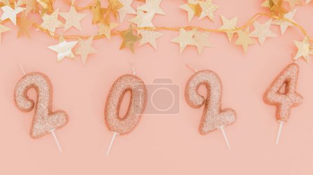 Photo for Shiny candles number 2024 with a festive gold garland in the shape of stars lie on a pink background, flat lay close-up. - Royalty Free Image