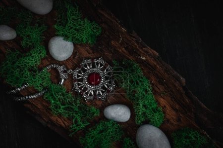 Foto de Un amuleto ritual con una joya roja, musgo verde y piedras grises en la corteza de un árbol se encuentra diagonalmente sobre un fondo negro, vista superior de cerca. Concepto esotérico, estilo oscuro. - Imagen libre de derechos