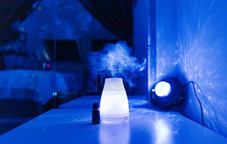 Foto de Un ambientador eléctrico con una botella de aceite aromático y una lámpara de noche se encuentran en una mesa en un salón de belleza en casa en luz de neón con vapor evaporador y reflexión en el espejo, vista lateral de cerca - Imagen libre de derechos