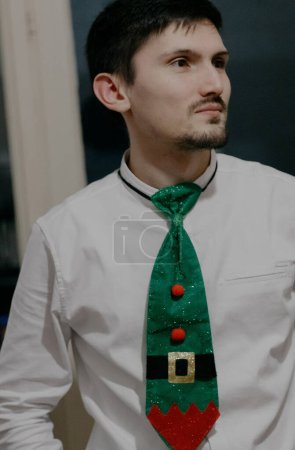 Photo for Portrait of one young handsome brunette guy in a white shirt with a green Christmas Santa Claus tie looking to the side, close-up side view. - Royalty Free Image