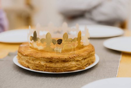 Téléchargez les photos : Une appétissante galette royale feuilletée avec une couronne en papier doré se trouve dans une assiette sur une table en bois, vue latérale rapprochée. - en image libre de droit