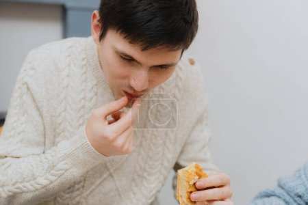 Téléchargez les photos : Un jeune beau mec caucasien sort joyeusement un cadeau surprise d'un morceau de galette royale de sa bouche, assis à une table dans la cuisine près du réfrigérateur, vue latérale rapprochée. - en image libre de droit