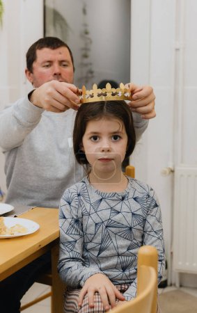 Téléchargez les photos : Un jeune homme blanc beau met une couronne de papier doré sur sa petite fille après avoir mangé une galette royale et trouvé un cadeau, assis à la table dans la cuisine, vue latérale rapprochée. - en image libre de droit