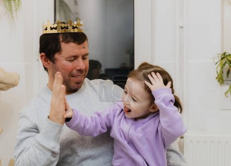 Téléchargez les photos : Un jeune homme blanc beau avec une couronne en papier doré sur la tête joue à applaudir sa petite fille après avoir mangé une galette royale et trouvé un cadeau, assis à la table dans la cuisine - en image libre de droit