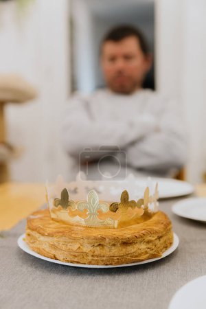 Téléchargez les photos : Une délicieuse galette royale feuilletée avec une couronne en papier doré se trouve dans une assiette sur une table en bois, sur le fond d'un homme flou, vue de côté gros plan. - en image libre de droit
