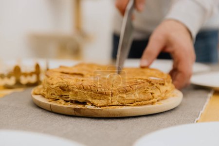One Caucasian young blurred man cutting into pieces a kings galette in a plate on a wooden board using a knife, holding with his hand, standing at the table, bottom close-up view with selective focus