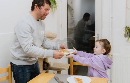 Téléchargez les photos : Une petite fille caucasienne prend une assiette avec un morceau de biscuit royal des mains d'un père heureux et souriant debout à la table dans la cuisine près de la porte vitrée avec accès à la cour arrière - en image libre de droit