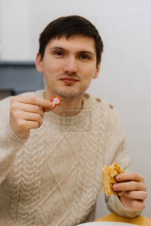 Téléchargez les photos : Un jeune beau mec caucasien avec un sourire montre un jouet surprise d'un morceau mangé de galette royale, assis à une table dans la cuisine, vue latérale rapprochée. - en image libre de droit