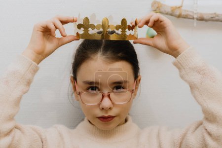 Téléchargez les photos : Une belle fille caucasienne avec des lunettes pose solennellement une couronne en papier doré sur sa tête, après avoir mangé une galette royale, assise à la table dans la cuisine, vue rapprochée d'en haut. - en image libre de droit