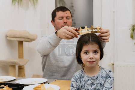 Téléchargez les photos : Un jeune homme blanc beau met une couronne de papier doré sur sa petite fille après avoir mangé une galette royale et trouvé un cadeau, assis à la table dans la cuisine, vue latérale rapprochée. - en image libre de droit
