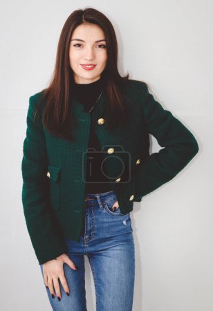 Foto de Retrato de una hermosa joven morena caucásica con ojos marrones, pelo largo y liso en una chaqueta verde de moda, vaqueros azules de pie con una sonrisa en la cara contra el fondo de un blanco - Imagen libre de derechos