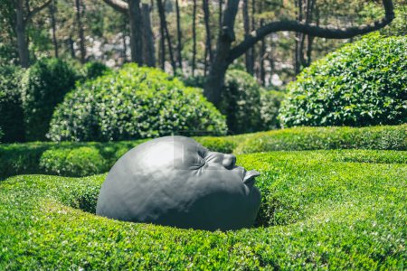 Foto de Una hermosa vista de una escultura de metal gris en forma de cabeza humana con una lengua sobresaliente en arbustos verdes en un día soleado de verano en el jardín botánico de maravillas en Francia, vista lateral de cerca. - Imagen libre de derechos