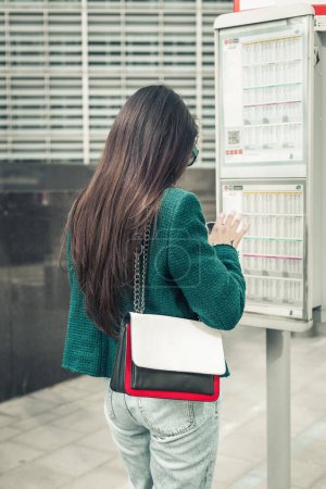 Téléchargez les photos : Une jeune fille brune caucasienne aux cheveux longs dans une veste verte se tient dos à un arrêt de bus près du tableau des horaires, vue latérale rapprochée. - en image libre de droit