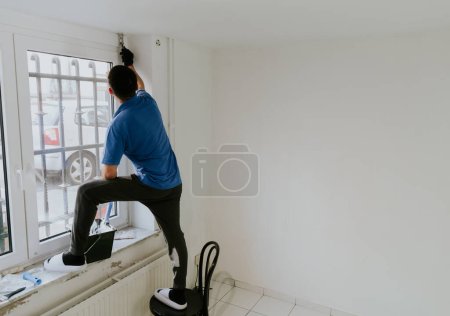 Téléchargez les photos : Un jeune homme caucasien barbu aux cheveux foncés et beau en T-shirt bleu se tient sur une chaise sur le côté près de la fenêtre et peint la fenêtre ouvrant avec un pinceau avec de la peinture blanche, vue latérale rapprochée avec - en image libre de droit