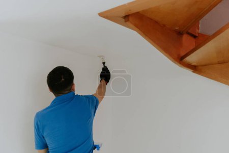 Téléchargez les photos : Un jeune homme caucasien aux beaux cheveux bruns dans un T-shirt bleu se tient du dos et peint avec un pinceau avec de la peinture blanche les coins du mur sous les escaliers en bois, vue latérale rapprochée avec - en image libre de droit