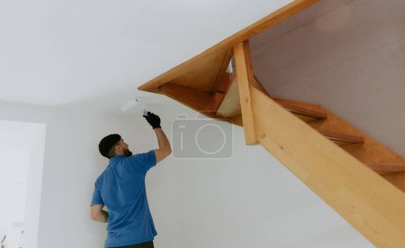Téléchargez les photos : Un jeune homme caucasien aux cheveux bruns et beau en t-shirt bleu se tient derrière et peint avec un rouleau le plafond sous un escalier en bois à côté d'une porte, vue rapprochée du bas avec - en image libre de droit