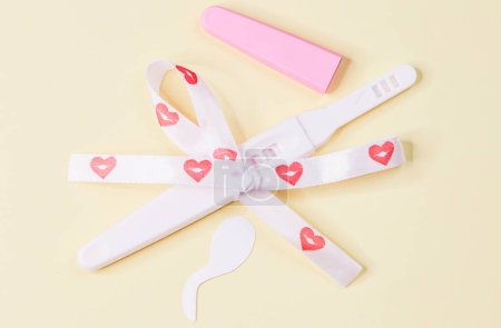 Photo for One pregnancy test tied with a ribbon with hearts in a bow and light yellow paper sperm on a background, close-up top view. - Royalty Free Image
