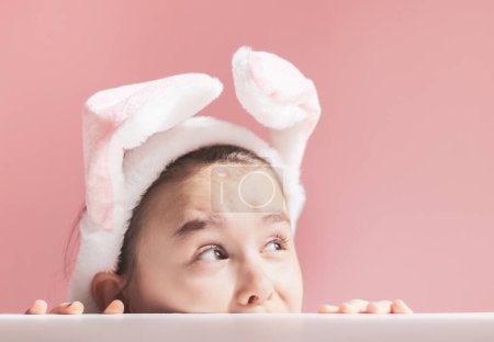 Beautiful cute caucasian girl peeking and looking to the side with headband pink and white easter bunny ears on white and pink background with little copy space ,side view closeup.Concept,banners