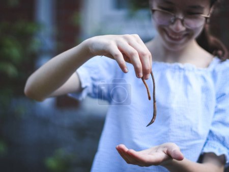 Photo for One beautiful Caucasian girl with a smile on her face holds an earthworm in her hands, standing in the garden at the backyard of a house on a spring morning, close-up side view with depth of field. - Royalty Free Image