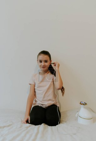 Eine schöne kaukasische Brünette mit gesammelten Haaren und in einem rosafarbenen T-Shirt behandelt ihr Ohr mit einem Infrarotlichtgerät, sitzt kniend auf einem Bett zu Hause in der Nähe einer weißen Wand, Nahaufnahme von der Seite.
