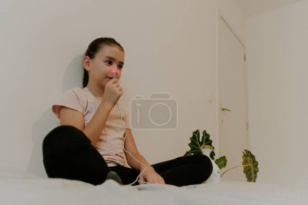 Foto de Una hermosa morena caucásica con el pelo recogido y en una camiseta rosa trata el pasaje nasal derecho con un aparato con luz infrarroja, sentado en la cama, con las piernas cruzadas, apoyado en una pared blanca, vista de cerca desde abajo. - Imagen libre de derechos